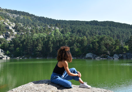 Young woman sitting on the edge of a mountain lake and looking into the distance. Black woman with curly afro hair sitting on top of a rock. Observing the Black Lagoon.のeditorial素材