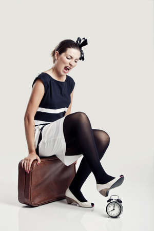 Beautiful young fashion woman sitting over a suitcase and smashing a clockの写真素材