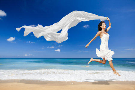 Beautiful woman running and jumping on the beach with a white scarfの写真素材