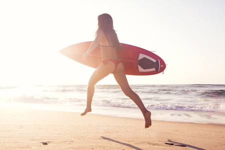 Hot surfer girl running in the beach with her surfboard at sunsetの写真素材