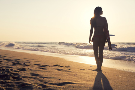 Beautiful surfer girl walking in the beach with her surfboard at sunset