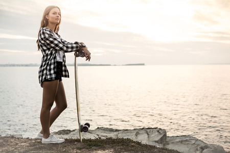 Beautiful and fashion young woman posing at the sunset with a skateboardの写真素材