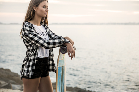 Beautiful and fashion young woman posing at the sunset with a skateboardの写真素材