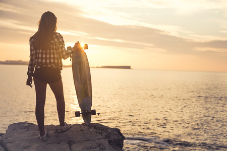 Beautiful and fashion young woman posing at the sunset with a skateboardの写真素材
