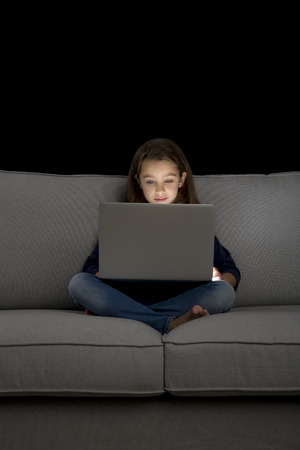 Happy little girl sitting on a couch and working a laptopの写真素材
