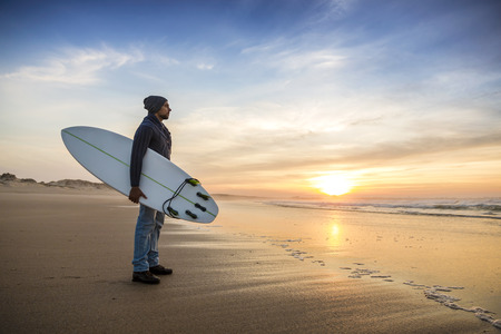 A surfer with his surfboard at the sunset looking to the waves