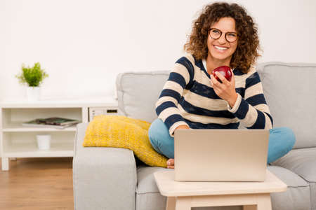 Beautiful woman at home sitting on the sofa working and eating a appleの写真素材