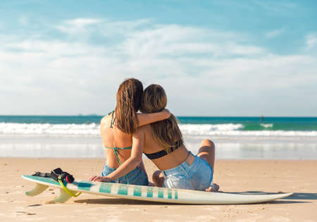 Two beautiful female friends at the beach sitting on the sand close to her surfboards embracing each otherの写真素材