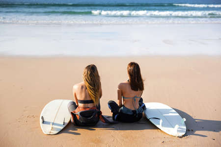 Two beautiful female friends at the beach sitting on the sand with her surfboards and looking to the wavesの写真素材