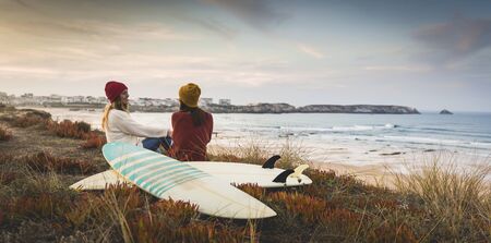 Two best friends sitting near the coastline with her surfboards while looking to the ocean
