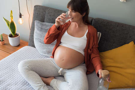 Pregnant woman holding glass of water relaxing in bed at homeの写真素材