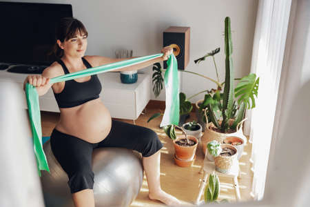Pregnant woman working out at home doing exercises on a fitball with a resistance band. Keeping in good shape while waiting for babyの写真素材