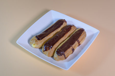 Three fresh eclairs on a white corner plate. On a beige background