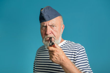 An old sailor with a smoking pipe. Portrait of a sailor In a marine vest striped shirt and a blue capの写真素材