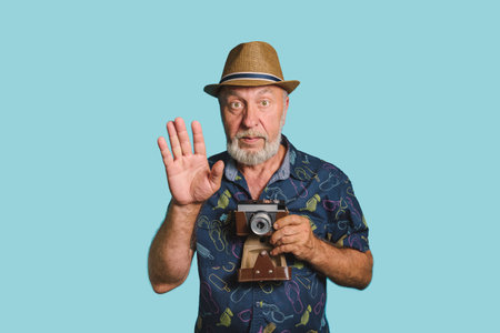Strike a pose. Eldery man with gray beard and straw hat holding old camera rise his hand up to take picture. Blue background portraitの写真素材