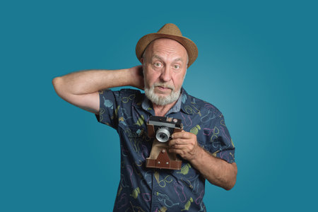 Senior photographer with retro camera scratching head. Mooved his straw hat back. frontal portrait dark blue backgroundの写真素材