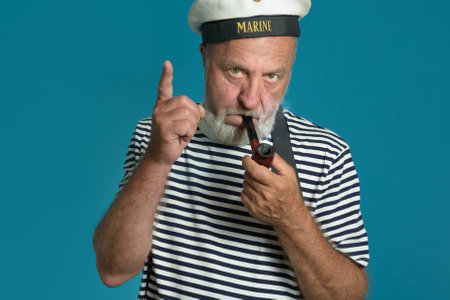 An old sailor smoking a pipe In a marine vest striped shirt and a marine cap - Portrait on blue backgroundの写真素材