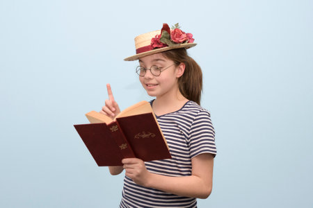 A girl in retro style clothes enthusiastically reads a book a straw hat with flowers, glasses and a book in her hand.の写真素材