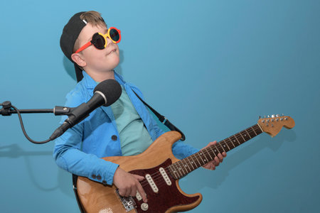A boy with a microphone in a baseball cap and sunglasses plays the electric guitar. On the blue. Place for textの写真素材