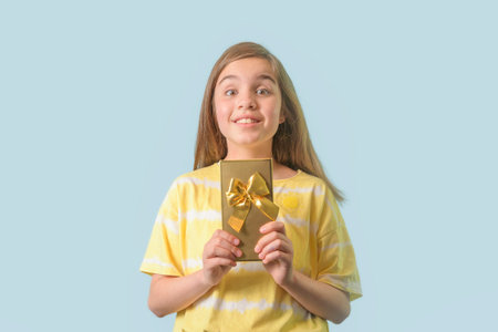 Joyful teenage girl - holds a gift in a gilded package with a bow in her hands. Light blue backgroundの写真素材