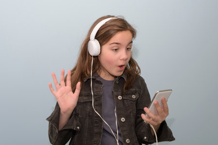 Teenage girl greets a friend with a white mobile phone and white headphones in the messenger. Light gray background. portraitの写真素材