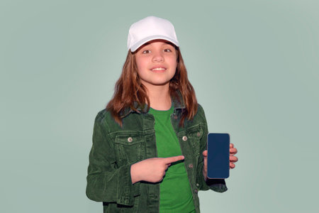 A teenage girl shows the screen of her smartphone. White baseball cap mocap, denim jacket and green shirt. light green backgroundの写真素材