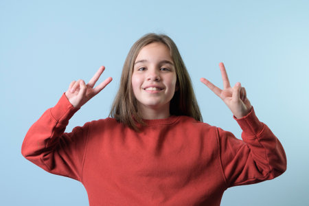 A teenage girl makes a victory sign with her fingers with both hands. Red sweater, blue backgroundの写真素材