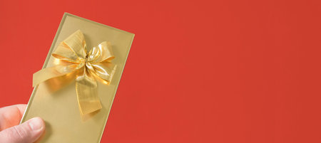 Banner - a man's hand with a gift, gold packaging in a festive bow. Red background. Copy spaceの写真素材