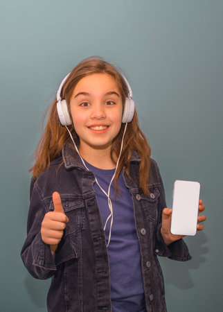 A girl is holding a white cell phone and smiling. She is wearing headphones and is giving a thumbs upの写真素材