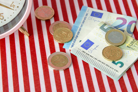 A pile of coins and a stack of euros sit on a red and white striped table and table clockの写真素材