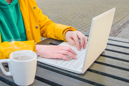A teenager sits outside with a white laptop and a cup of coffee