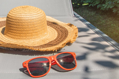 A straw hat and sunglasses are on a patio chair. The hat is brown and white, and the sunglasses are...