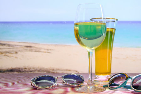The scene is set on a beach, with the ocean in the background. A glass of wine and a glass of beer sit on a table next to a pair of sunglasses. Scene is relaxed and leisurelyの写真素材