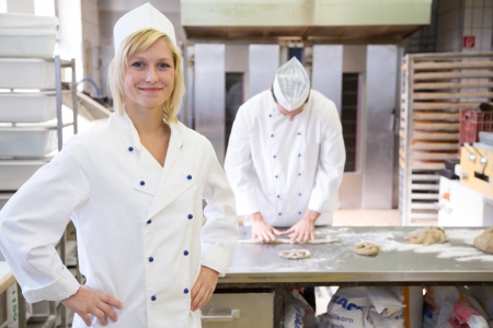 Baker posing in bakery or bakehouse. Another baker is working in the background