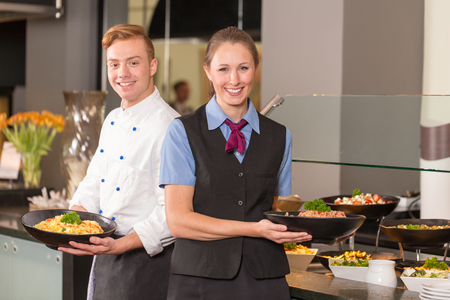 cook or chef and waitress from catering service posing with food in front of buffetの写真素材