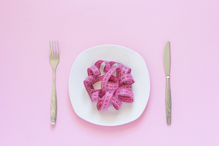 Pink measuring tape lying on plate in the form of spaghetti, knife and fork on pink background. Diet or anorexia concept. Top viewの写真素材