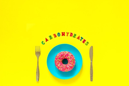 Pink donut on blue plate and cutlery table knife fork Text Carbohydrates. Still life on yellow background. Copy space Top view Flat lay Concept No Diet dayの写真素材