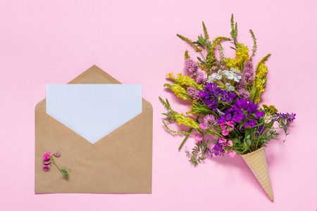 Bouquet of field multicolored flowers in waffle cone and craft envelope with white empty paper card for text on pink background. Greeting card Flat Lay Mock up Concept Women's day or Mothers Day.の写真素材