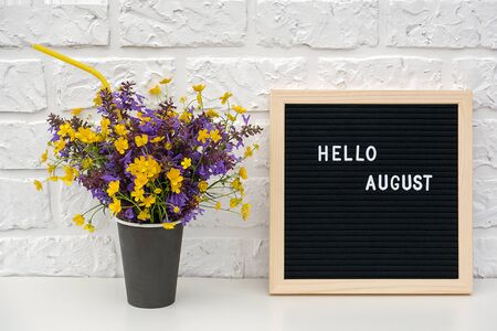 text Hello August on black letter board and bouquet of colored flowers in black paper coffee cup with cocktail straw on background white brick wall. Template Mock up Concept.の写真素材