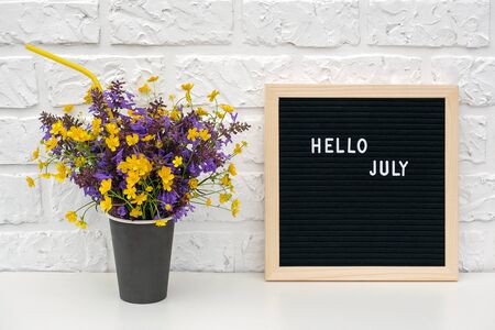 text Hello July on black letter board and bouquet of colored flowers in black paper coffee cup with cocktail straw on background white brick wall. Template Mock up Concept.の写真素材