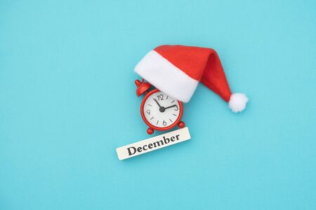 Wooden calendar winter month December, red alarm clock and Santa hat on blue paper background. Concept Hello December, xmas. Creative Top view Flat Lay.の写真素材