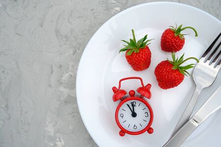 Red ripe strawberries berry on white plate, cutlery and red alarm clock on gray stone background table, close up. Top view, flat lay, copy space. Concept diet and detox time or summer menu time.の写真素材