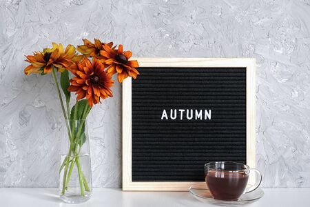 Autumn text on black letter board, bouquet of orange flowers in vase and cup of tea on table against grey stone wall. Template for postcard, greeting card Concept Hello autumn.の写真素材
