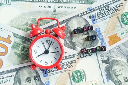 Time is money text and red alarm clock on background of American hundreds dollar bills, closeup....