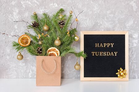Happy Tuesday text on black letter board and festive bouquet of fir branches with christmas decor in craft package on table. Template for postcard, greeting card Concept Hello winter Tuesday.の写真素材