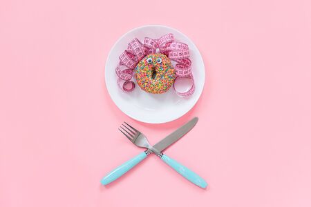 Abstract funny face of woman from donut with eyes and hair from centimeter tape on plate, cutlery on pink background. Diet or unhealthy food concept Top view Flat lay Copy space.の写真素材