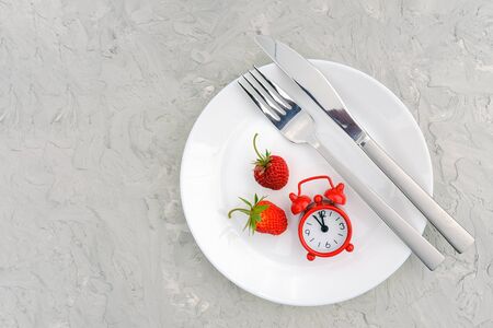 Red ripe strawberries berry on white plate, cutlery and red alarm clock on gray stone background table. Top view, flat lay, copy space Template. Concept diet and detox time or summer menu time.の写真素材