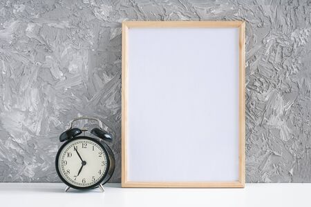 Wooden vertical white empty frame and black alarm clock on table on gray concrete wall background. Mockup Template for your design.の写真素材