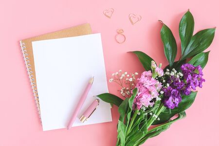 Blank white sheet on spiral golden notepad with paper clip heart and ring and bouquet of flowers on pink background. Concept Romantic love Mockup for your text and design. Top view Flat lay .の写真素材