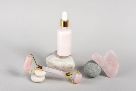 Crystal rose quartz facial roller, massage tool Gua sha and anti-aging collagen, serum in glass bottle on stones, grey background. Facial massage for natural lifting, Beauty concept.の写真素材